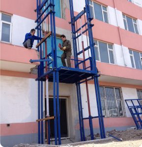 Two workers are working on a blue freight elevator exterior a building.