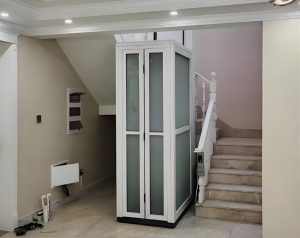 A white home lift with glass panels and a white cabin frame located between stairs in a home.